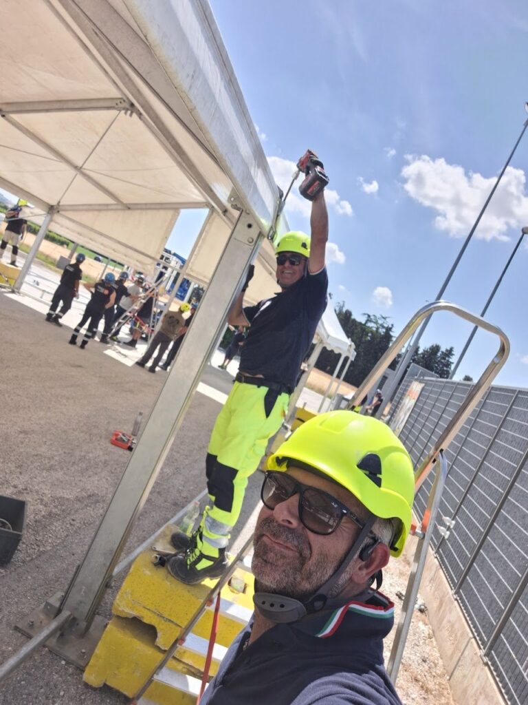 Due volontari con casco giallo al lavoro sulla struttura di una tenda, uno in alto con l'avvitatore