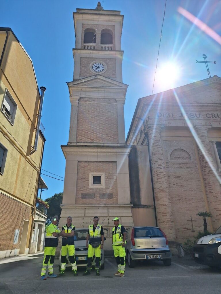 Quattro volontari del Gruppo CB Tigrotto in divisa gialla davanti al campanile della chiesa del SS. Crocifisso