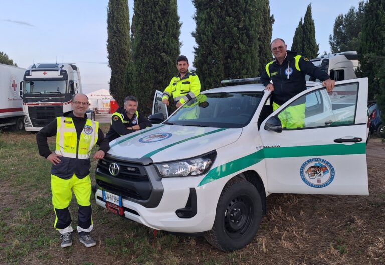 Quattro volontari accanto al pickup bianco dell'associazione, parcheggiato al campo base fra i camion della Croce Rossa