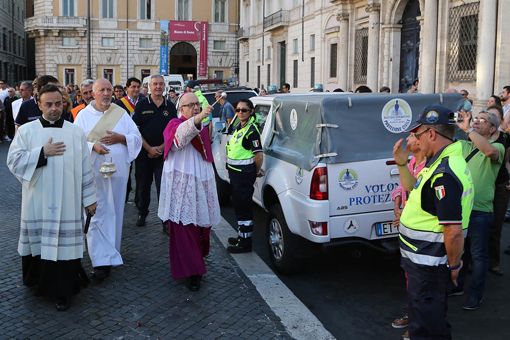 Un sacerdote benedice con l'aspersorio un pick-up della protezione civile in una piazza di Roma, davanti a un gruppo di volontari in divisa ad alta visibilità