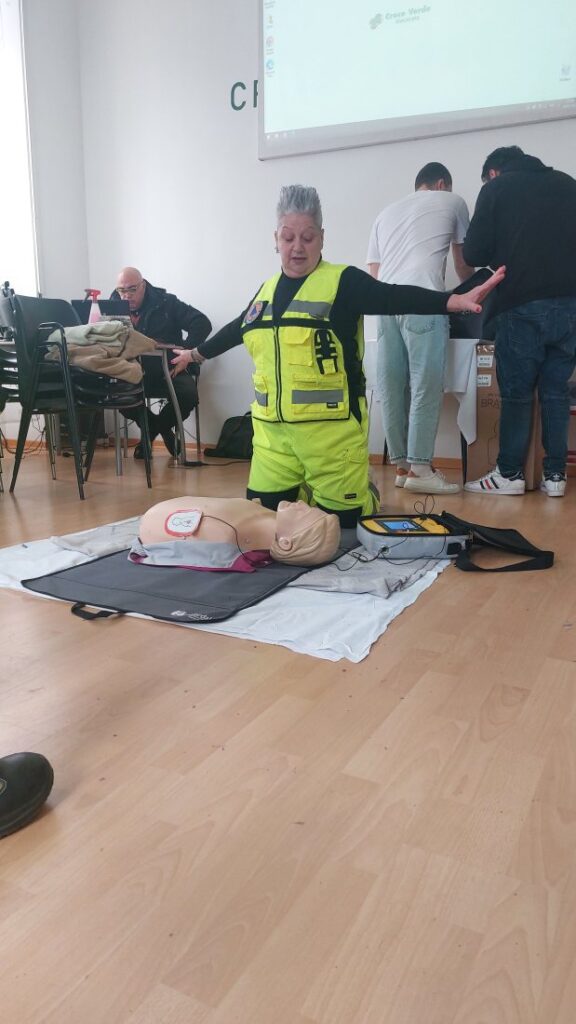 Un'istruttrice inginocchiata accanto al manichino allarga le braccia per far allontanare i presenti; sul torace del manichino sono applicate le piastre del defibrillatore