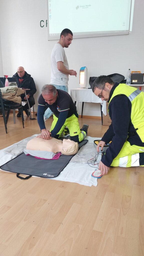 Due volontari lavorano insieme sul manichino: uno esegue le compressioni, l'altro applica le piastre del defibrillatore semiautomatico appoggiato a terra