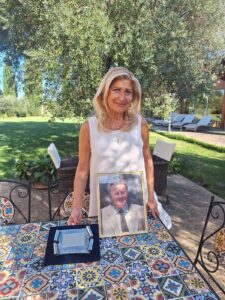 Cristina Emiliozzi in giardino, dietro un tavolo su cui sono appoggiati il ritratto del padre e la targa del 1989