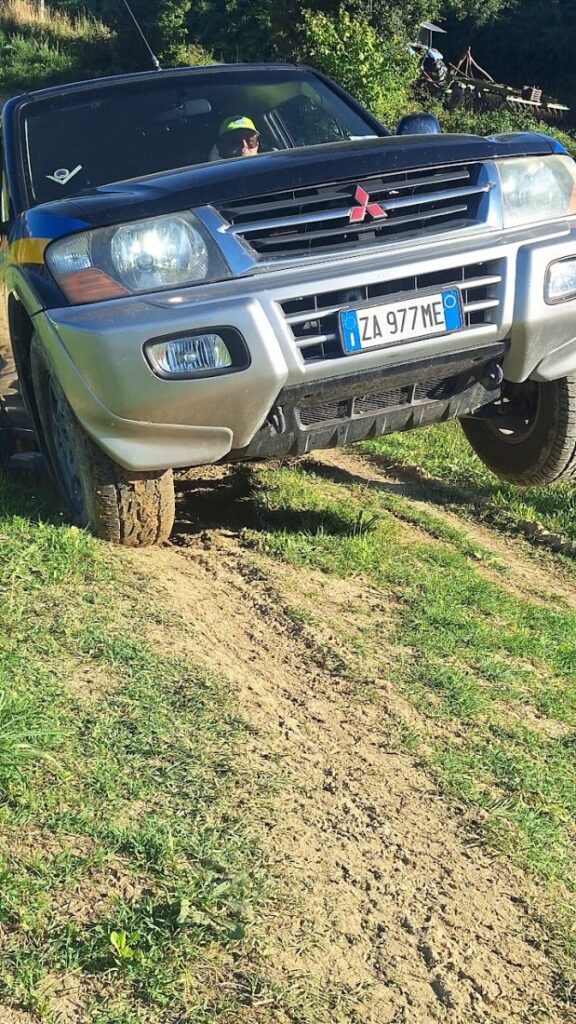Primo piano del muso del Pajero con la ruota anteriore destra sollevata sul bordo di un solco profondo, il conducente con il berretto giallo al volante