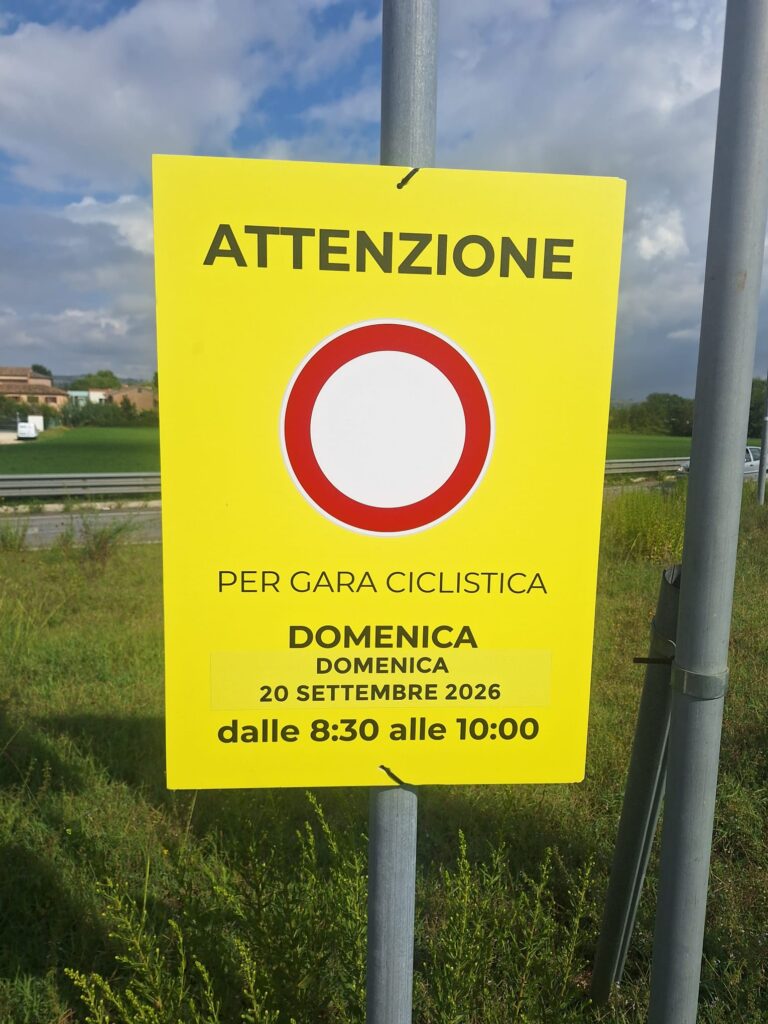 Cartello giallo legato a un palo lungo la strada: «Attenzione — divieto di transito per gara ciclistica — domenica 20 settembre 2026 dalle 8:30 alle 10:00»