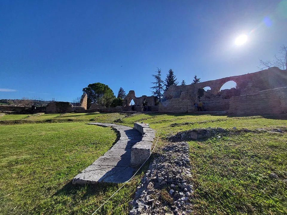 L'area archeologica di Helvia Recina in una giornata di sole: il prato dell'orchestra, i blocchi di pietra del proscenio e sullo sfondo le arcate del teatro