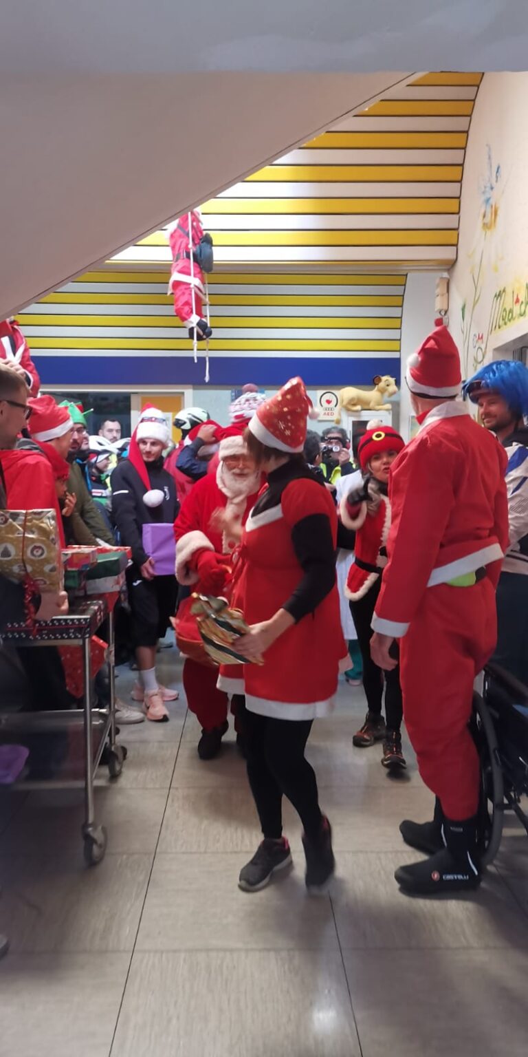 Dentro il reparto, un carrello carico di pacchi regalo e una persona vestita di rosso che ne porta uno; intorno, i partecipanti alla pedalata e il personale