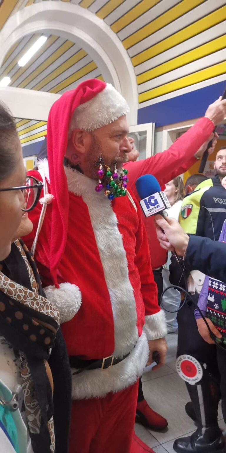 Un partecipante vestito da Babbo Natale, con le palline di Natale appese alla barba, intervistato da una troupe all'ingresso dell'ospedale; sullo sfondo un agente della Polizia Locale