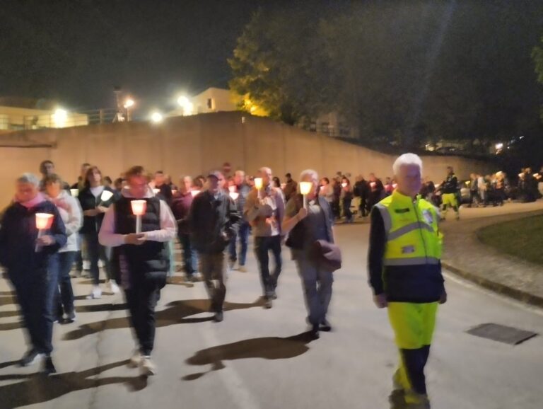 I pellegrini camminano di notte lungo la strada tenendo in mano le fiaccole accese dentro i bicchieri di carta; a destra, in divisa ad alta visibilità, un volontario accompagna la colonna