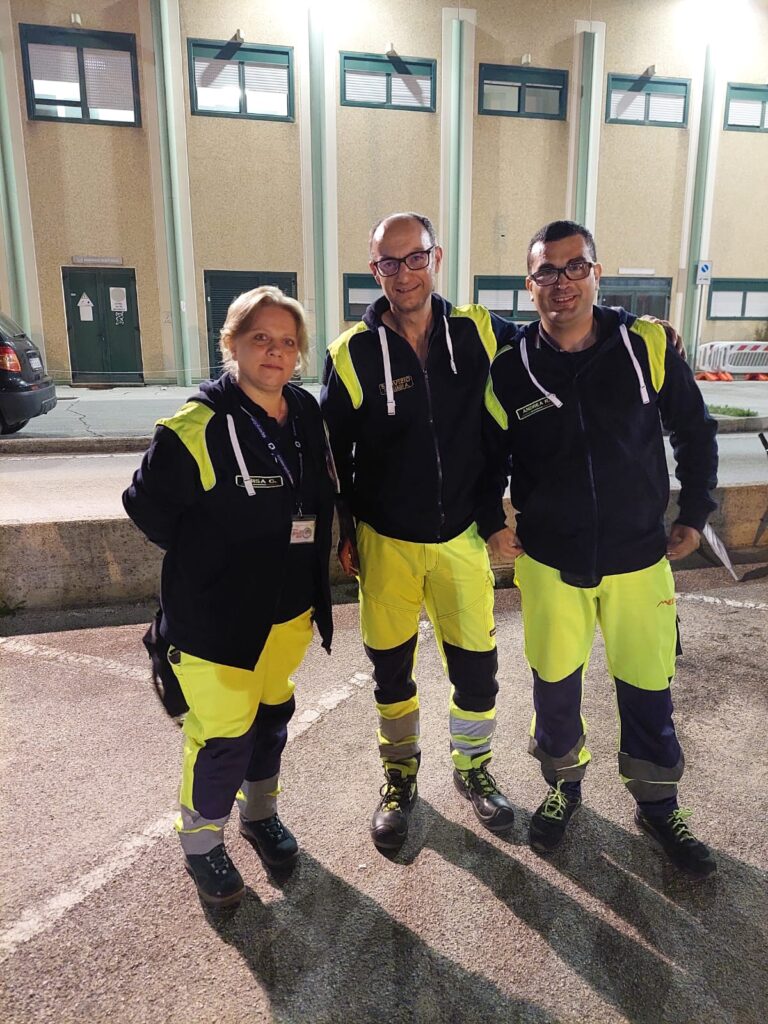 Tre volontari del Gruppo CB Tigrotto in divisa posano sorridenti nel piazzale dell'ospedale di Camerino, di sera, prima della partenza del pellegrinaggio