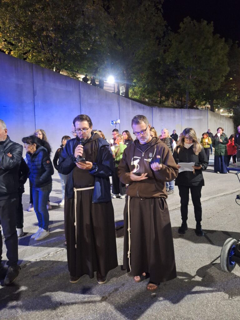 Due frati cappuccini in saio guidano la preghiera al microfono prima della partenza; dietro di loro una tromba della fonia mobile su un'asta e la gente con le candele