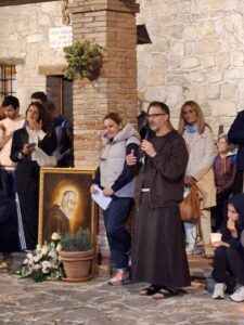 Un frate cappuccino parla al microfono accanto al ritratto di Padre Pio circondato da rose bianche, nel cortile del convento