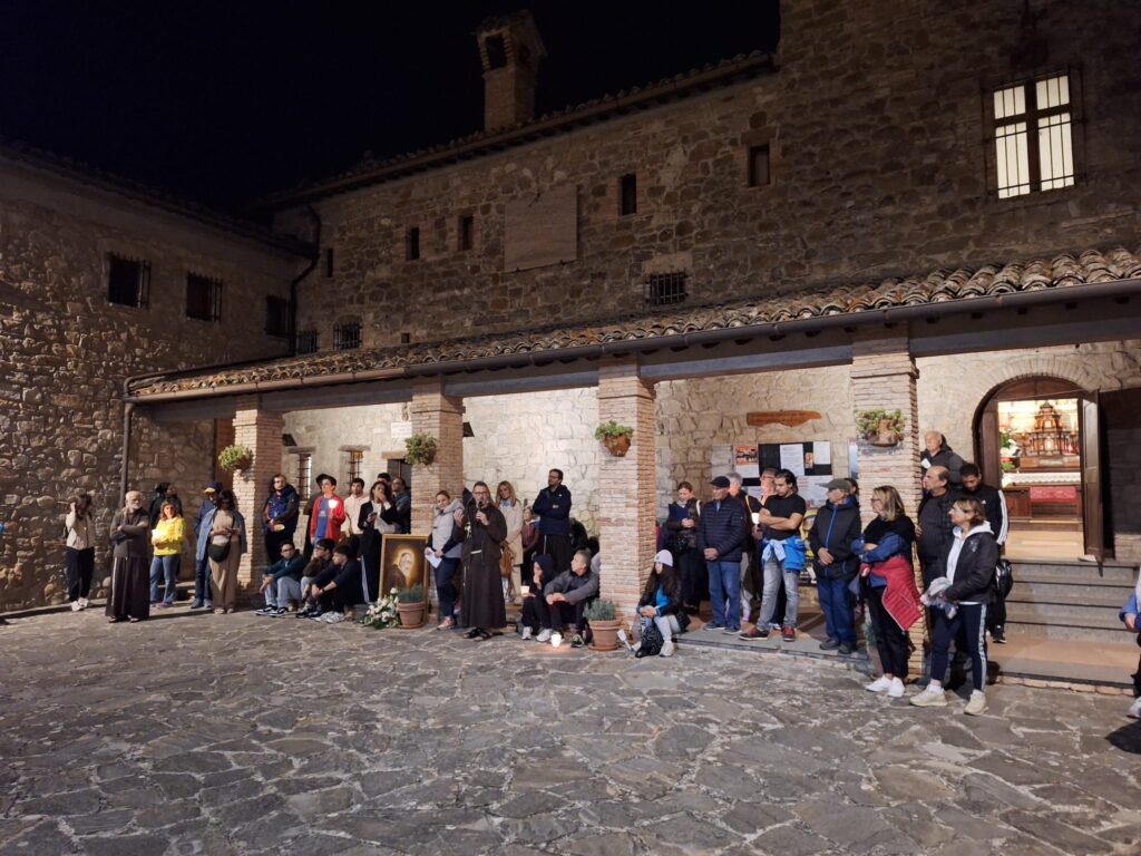 Il cortile del convento di Renacavata di notte, con la gente raccolta sotto il portico illuminato e il ritratto di Padre Pio appoggiato a terra con i fiori davanti