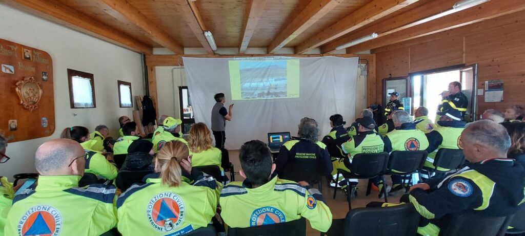 Una sala con soffitto in legno piena di volontari seduti in divisa gialla, che guardano una proiezione con la fotografia di un campo tende e la scritta «esempio di campo con tende (non ottimale)»