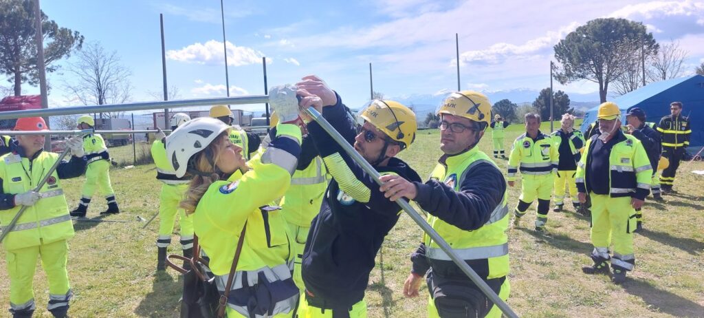 Volontari con caschi gialli e bianchi sollevano insieme i pali metallici del telaio di una tenda; sullo sfondo la catena dei Sibillini ancora innevata