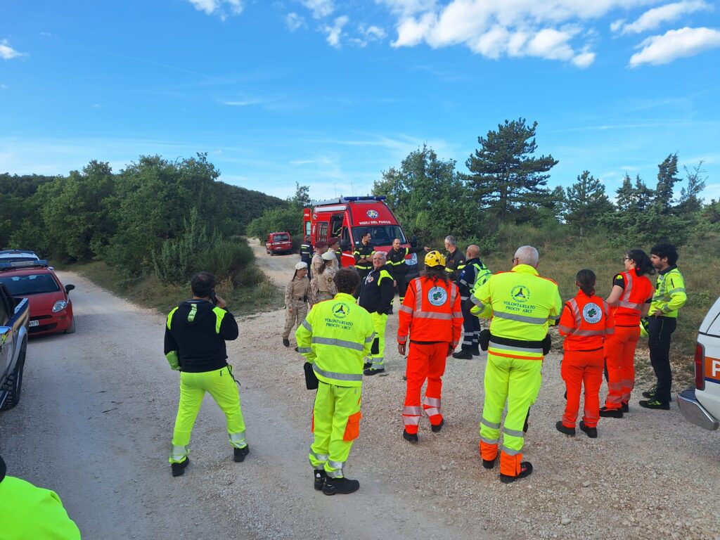 Un gruppo di volontari in divisa gialla e arancione, vigili del fuoco e militari dell'Esercito ascoltano le istruzioni in cerchio su una strada sterrata