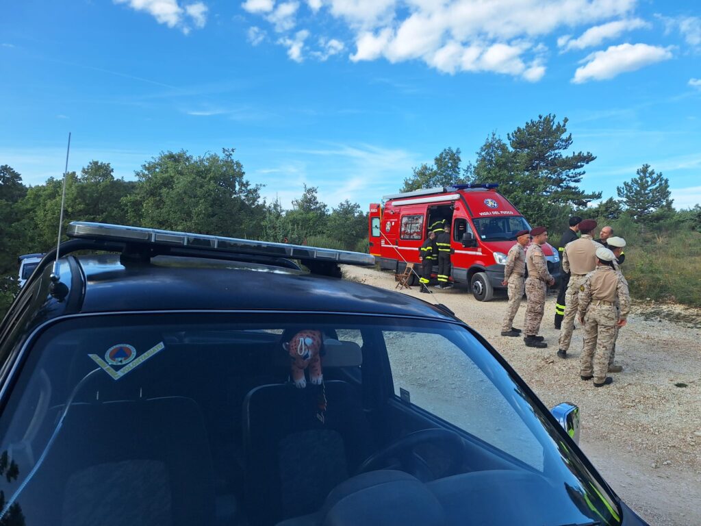 Un fuoristrada con l'adesivo della Protezione Civile sul parabrezza in primo piano; dietro, il furgone dei vigili del fuoco e un gruppo di militari