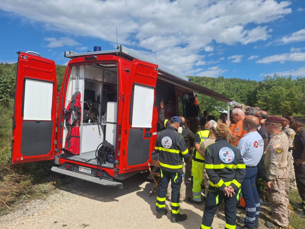 Il furgone comando dei vigili del fuoco con le porte posteriori aperte e il tendalino steso, circondato da volontari e militari che ascoltano