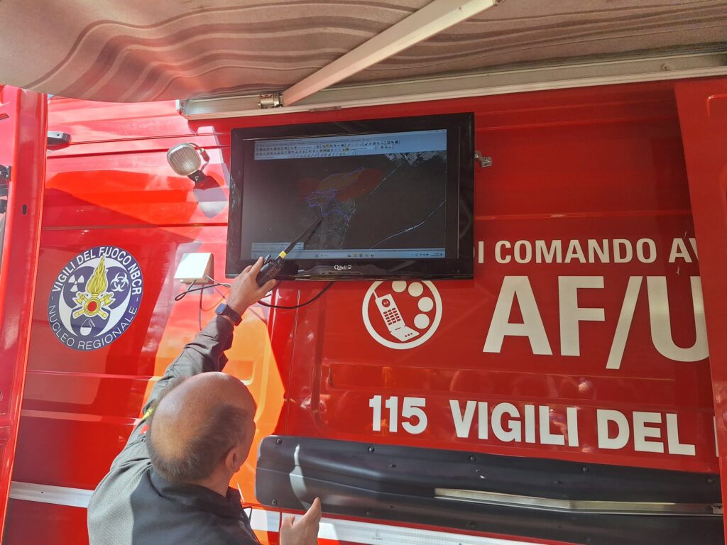 Un operatore indica con l'antenna della radio lo schermo montato sulla fiancata del furgone comando, dove una mappa mostra l'area di ricerca divisa in settori colorati