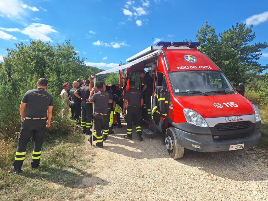 Vigili del fuoco in divisa radunati attorno al furgone comando sotto il tendalino, con il bosco alle spalle e il cielo sereno