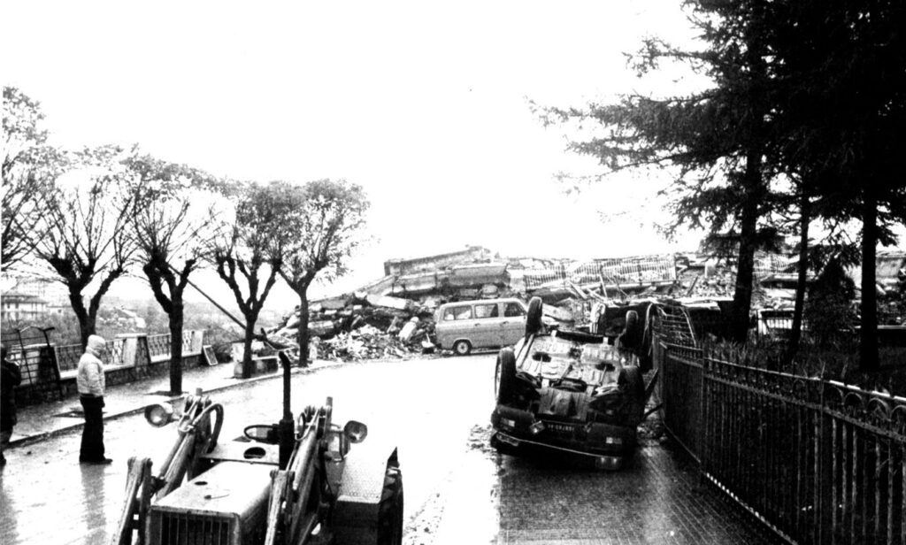 Una strada di Sant'Angelo dei Lombardi dopo il terremoto del 1980: un'auto ribaltata, palazzi crollati e una persona sola in piedi sul marciapiede