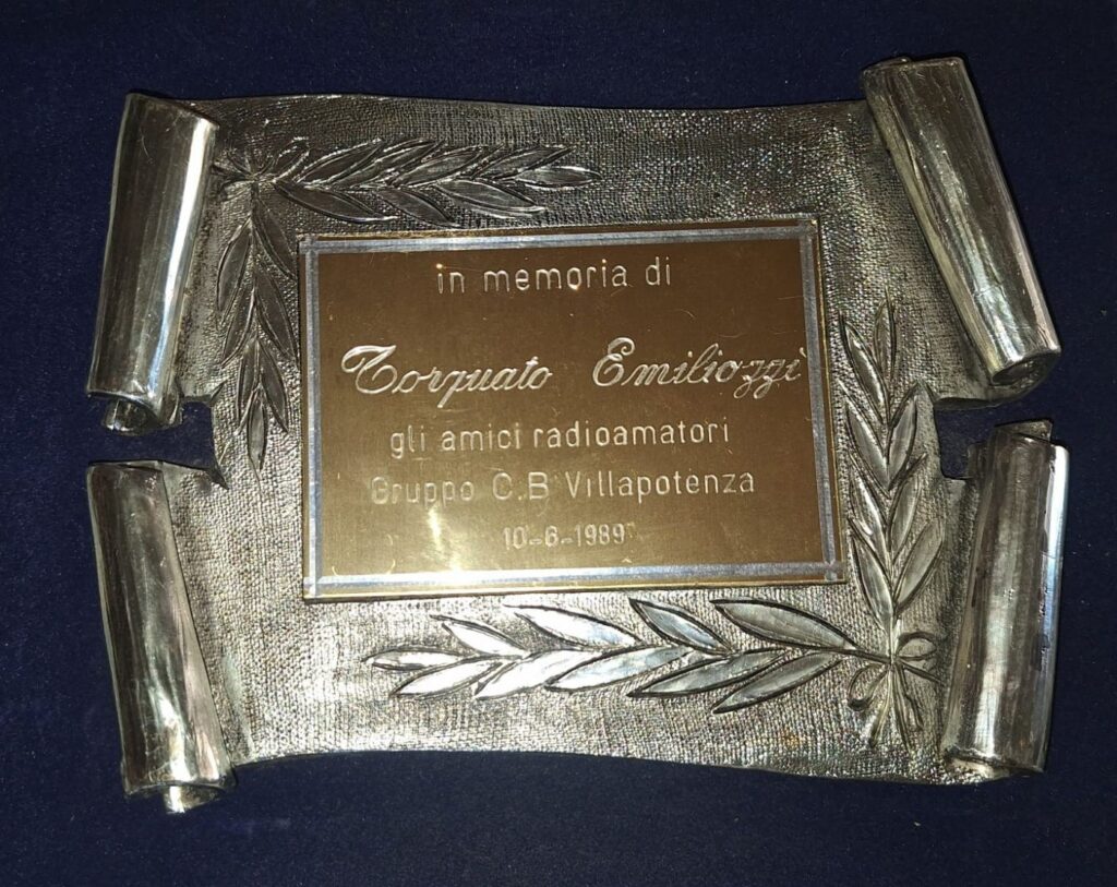Targa in metallo a forma di pergamena con la scritta «in memoria di Torquato Emiliozzi — gli amici radioamatori Gruppo C.B Villapotenza — 10-6-1989»