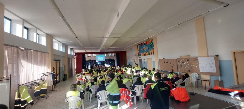 Una sala lunga piena di volontari seduti in divisa gialla che seguono una proiezione su uno schermo in fondo