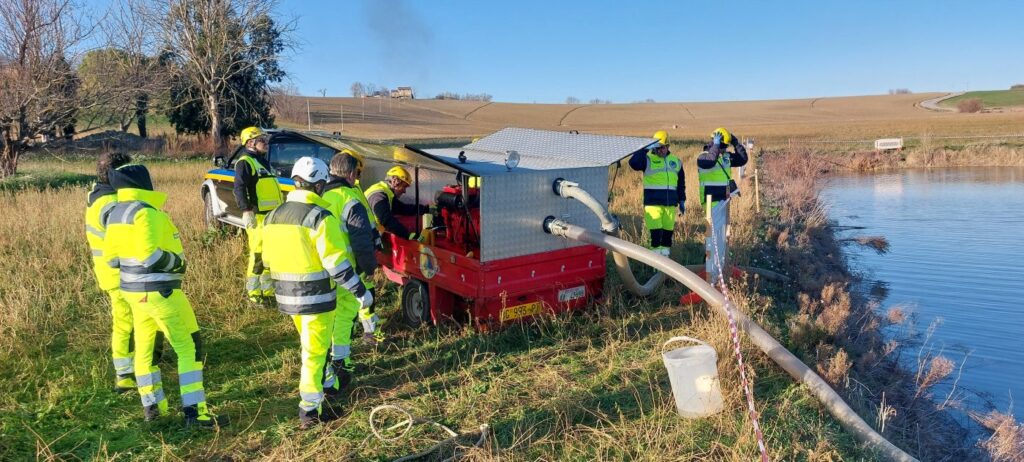 Una motopompa idrovora su carrello accesa in riva al laghetto, con il tubo di aspirazione immerso nell'acqua e alcuni volontari con il casco che la seguono da vicino